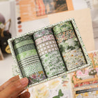 Hand holding Floral Whisper green forest washi tape set, showcasing the compact box and assorted roll widths against a floral background