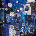 Variant image of the Celestial Ocean style Premium Mixed Media Junk Journal Kit in its blue and holographic packaging. Shows the assorted 30 pieces including the large laser-cut frame, the 'ice crystal' holographic PET butterflies and gears, and textured specialty paper through the clear window.