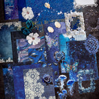 Variant image of the Celestial Ocean style Premium Mixed Media Junk Journal Kit in its blue and holographic packaging. Shows the assorted 30 pieces including the large laser-cut frame, the 'ice crystal' holographic PET butterflies and gears, and textured specialty paper through the clear window.