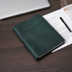 Dark green crazy horse leather journal closed on wooden desk with manuscript pages and fountain pen