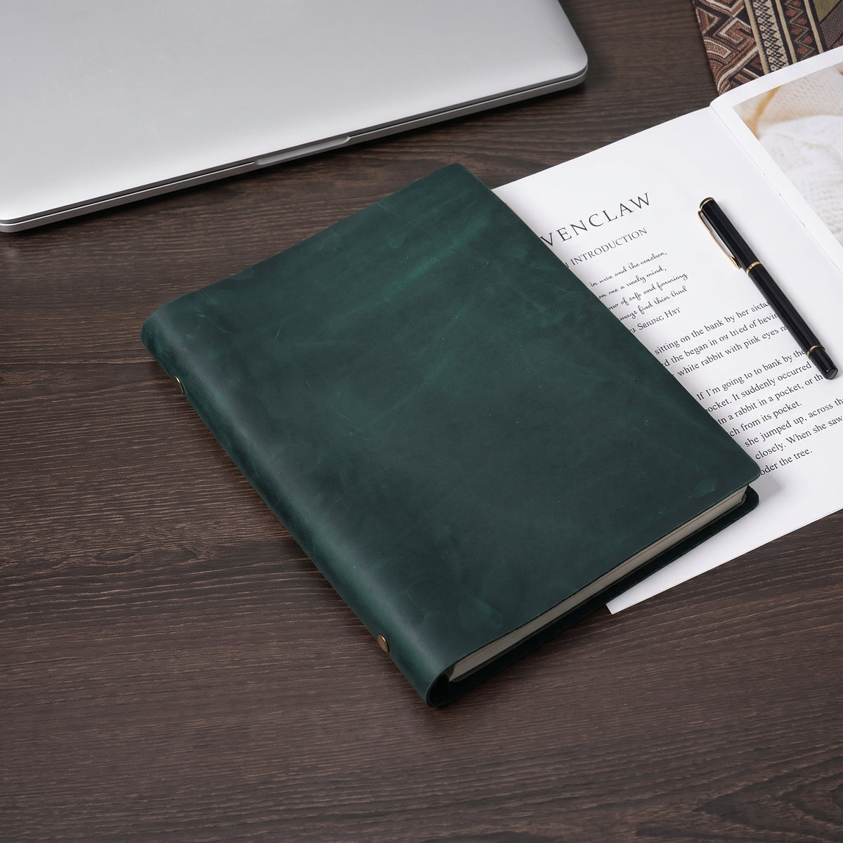 Dark green crazy horse leather journal closed on wooden desk with manuscript pages and fountain pen