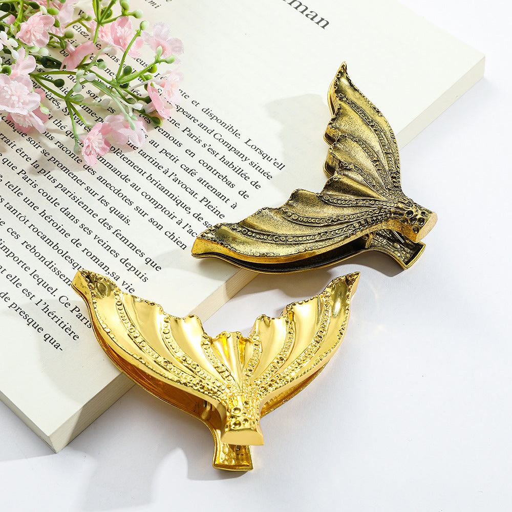 Luminous Siren Gold and Aged Antique Brass mermaid tail heavy-duty metal clips resting on an open vintage French book with delicate pink flowers, perfect for aesthetic junk journaling.