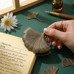 Detailed close-up of a hand securing an antique bronze ginkgo leaf journal clip onto a memo notebook page, showcasing its sturdy construction and vintage metallic finish.