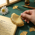 Close-up of a hand holding a vintage gold ginkgo leaf metal clip, demonstrating its heavy-duty clamping force on a thick notebook page for aesthetic planner organization.