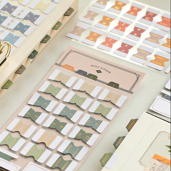 Vintage index sticker set on packaging card with numbered tabs and gold foil accents, arranged beside an open journal.