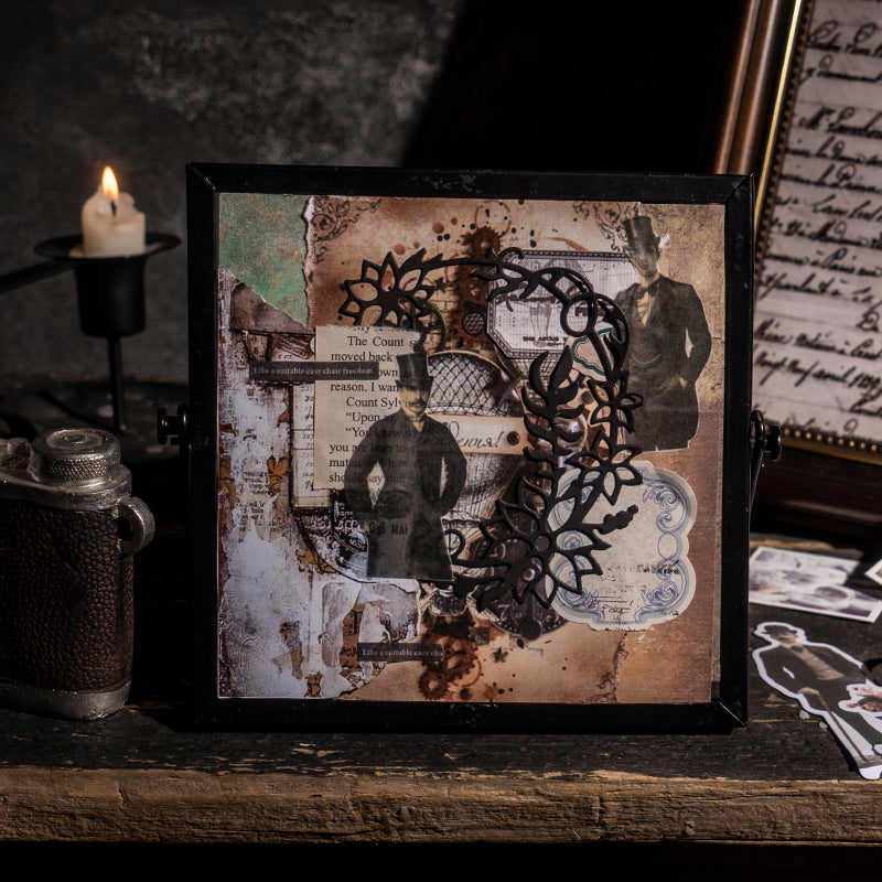  A framed junk journal collage artwork featuring vintage character stickers, ornate black lace cutouts, and distressed paper layers, displayed beside a lit candle and antique camera.