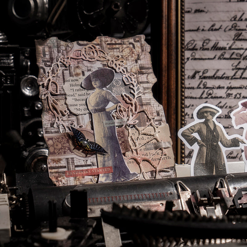 A detailed collage card leaning against a vintage typewriter, layered with a woman in a wide-brimmed hat, butterfly stickers, and ornate lace filigree.
