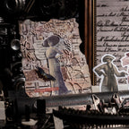 A detailed collage card leaning against a vintage typewriter, layered with a woman in a wide-brimmed hat, butterfly stickers, and ornate lace filigree.