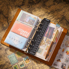 Open A5 crazy horse leather ring binder showing 6-ring mechanism, card slots, ruler insert, and fountain pen on vintage map paper.