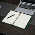 Open green leather ring binder journal with fountain pen beside a laptop on dark wood desk