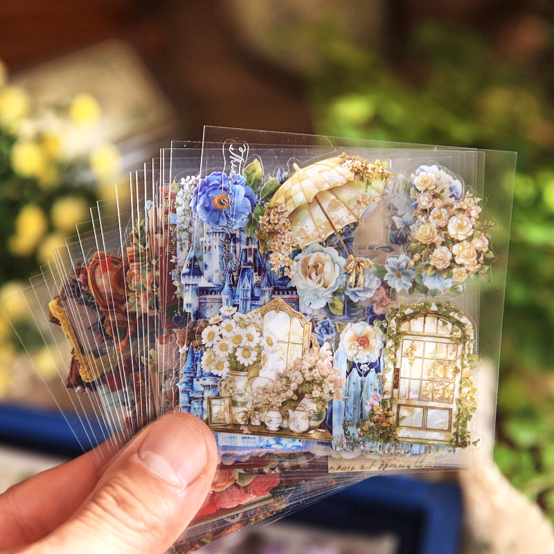 Hands holding open vintage world sticker gift box showing assorted sticker sheets with global landmarks, floral arrangements, and architectural designs inside