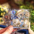 Hands holding open vintage world sticker gift box showing assorted sticker sheets with global landmarks, floral arrangements, and architectural designs inside