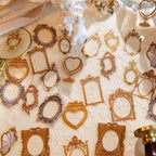 Assorted vintage gold frame PET stickers in round, heart, and crown shapes scattered on white lace with a small cherub figurine.