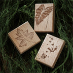 Three vintage beech wood rubber stamps displayed on dark green foliage background, carved botanical and leaf designs on smooth wooden blocks