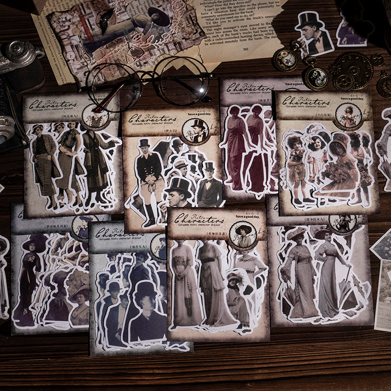  Eight vintage character sticker packs arranged on a wooden desk with antique glasses, pocket watches, and aged newspaper pages. Each pack displays die-cut sepia portraits in Victorian and Edwardian dress.