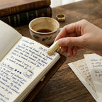 A close-up of a hand placing a solid metal cicada-shaped journal clip onto the open pages of a vintage leather notebook. The notebook features handwritten daily musings and a coffee cup doodle, set on a rustic wooden desk with a cup of coffee and antique books in the background.
