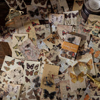  Overhead view of butterfly specimen vintage papers scattered across a dark wooden desk beside a brass magnifying glass and bulldog clip — dozens of illustrated moth and butterfly sheets from the Butterfly Specimens theme