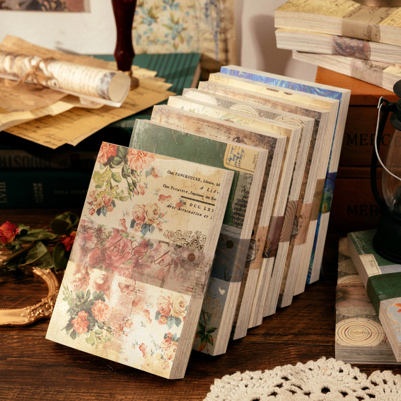 Eight vintage scrapbook paper pads displayed upright on a wooden desk, showing floral and manuscript cover designs with lace doilies.