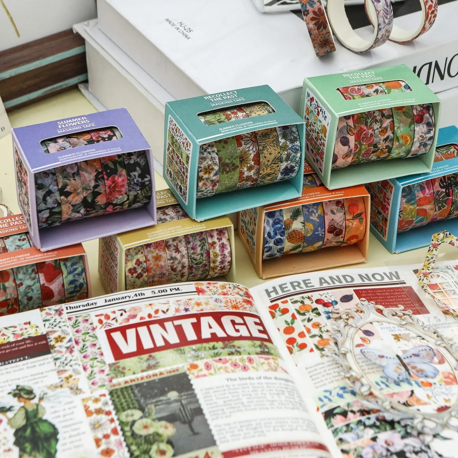 Several boxes of Summer Flowers botanical washi tape stacked elegantly behind an open vintage journal. Showcases the colorful packaging and aesthetic scrapbooking inspiration.