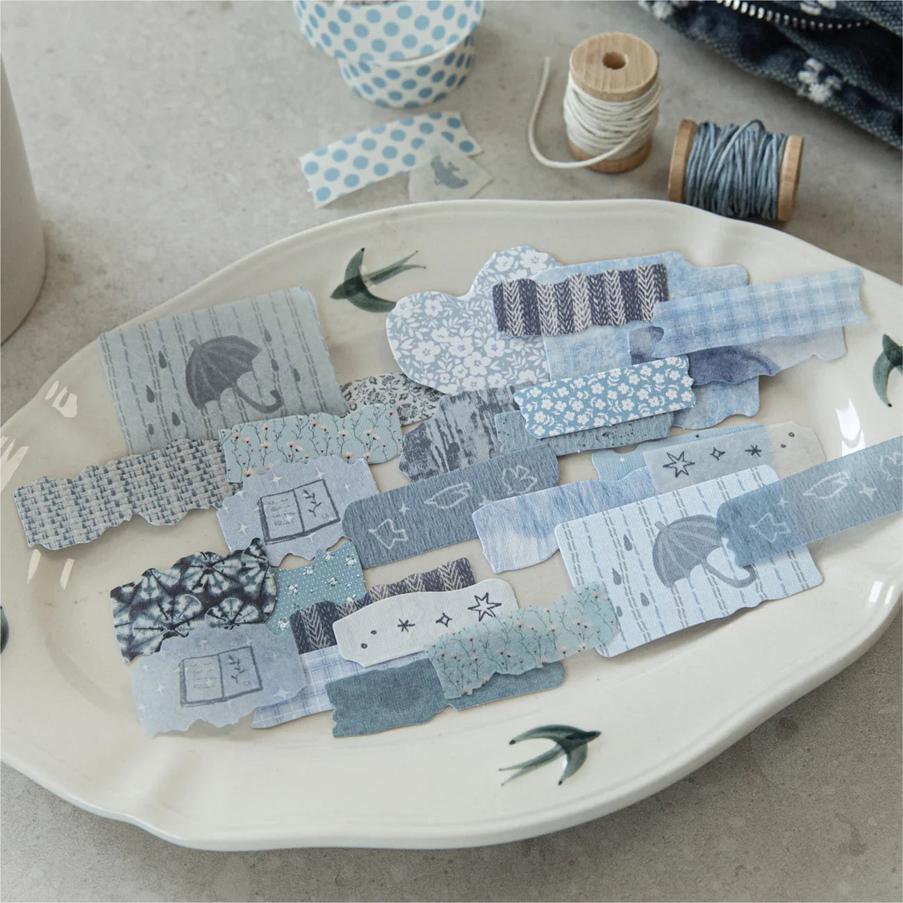 An elegant arrangement of Blue Overture faux-fabric stickers displayed on a vintage ceramic plate, featuring blue aesthetic elements like umbrellas and stars for creative bullet journaling.