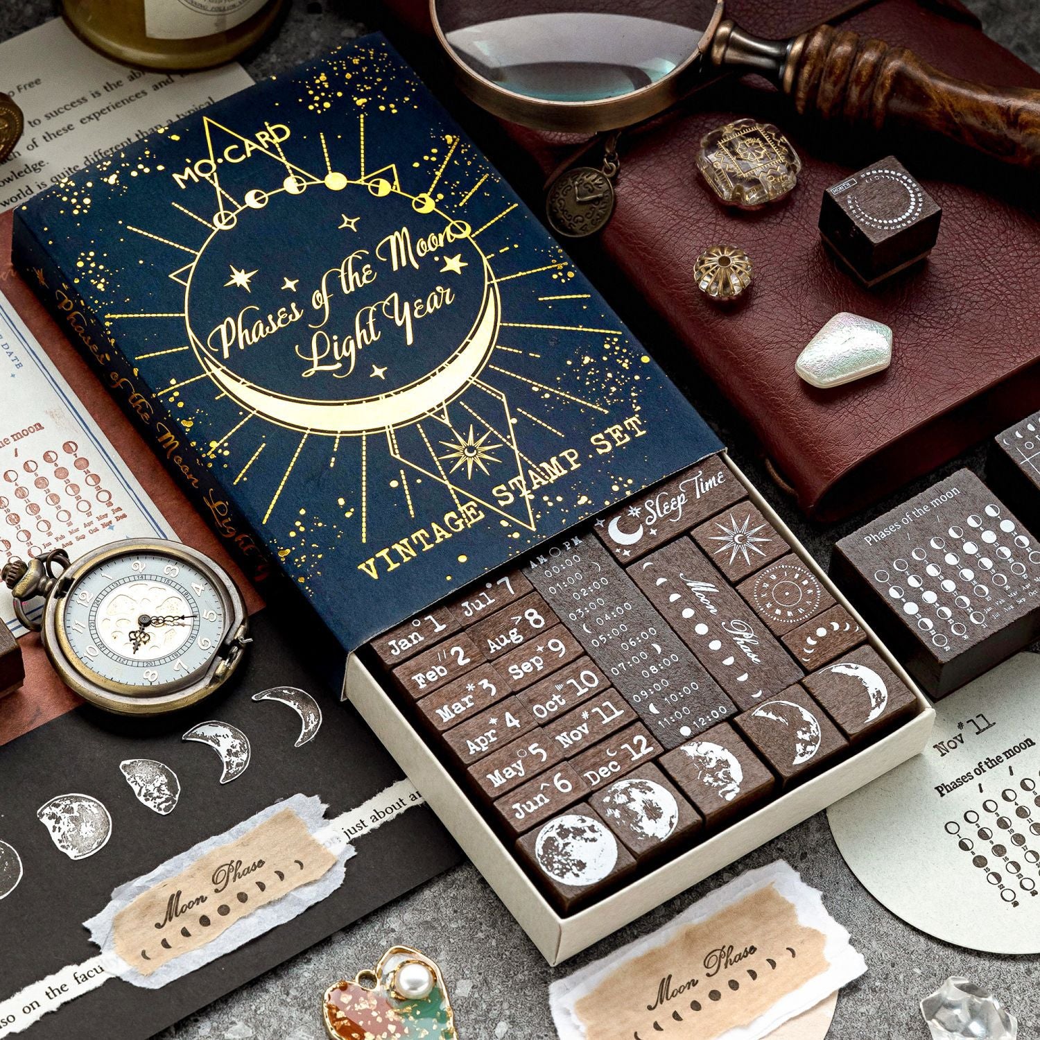 The Phases of the Moon celestial wooden stamp set partially opened in its elegant navy blue and gold-foil matchbox packaging. Styled with dark academia props like a vintage pocket watch and a magnifying glass.