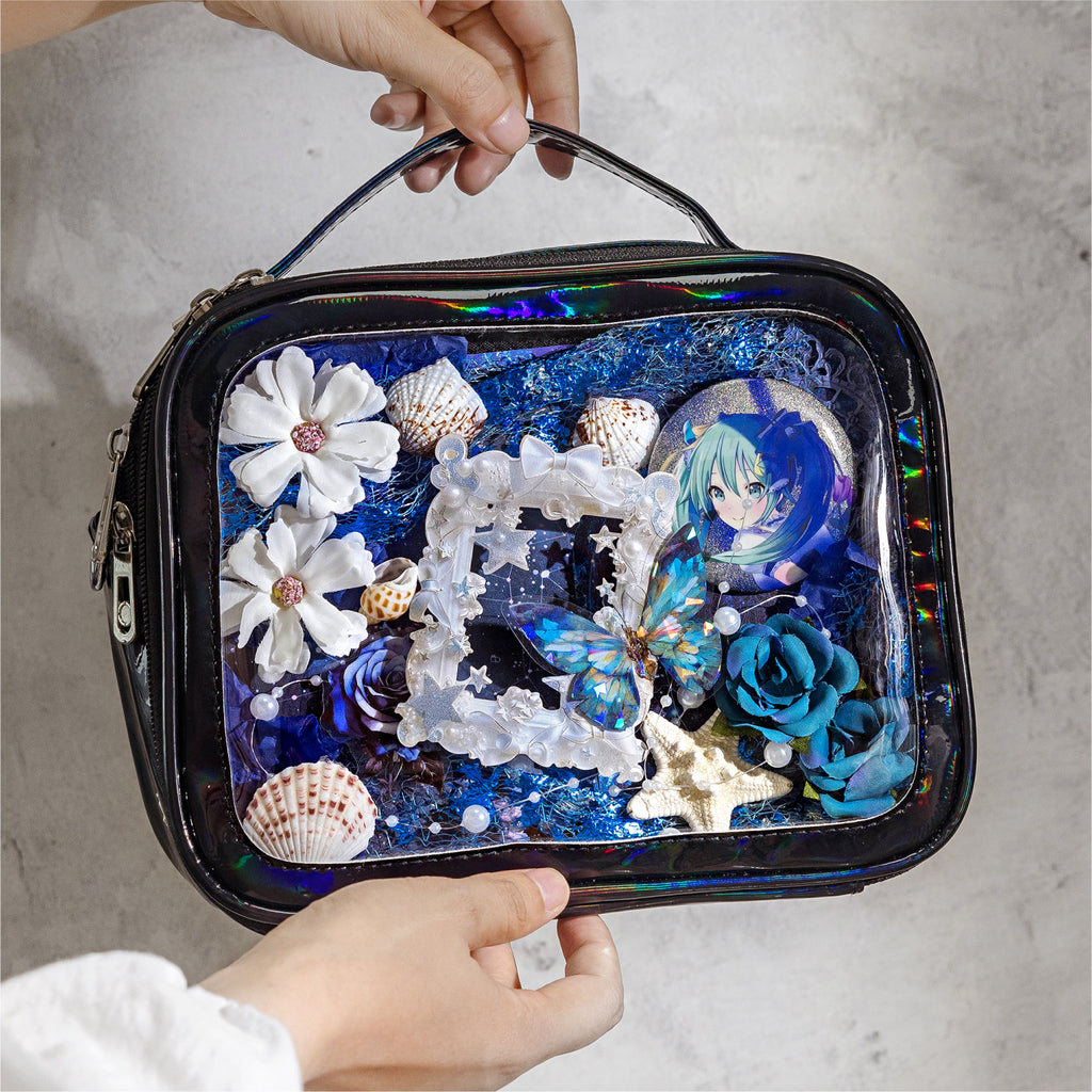 A person holding a black Ita bag decorated with the Celestial Ocean mixed media kit. The display window features an anime pin surrounded by blue mesh, white and blue 3D flowers, seashells, and a holographic butterfly.