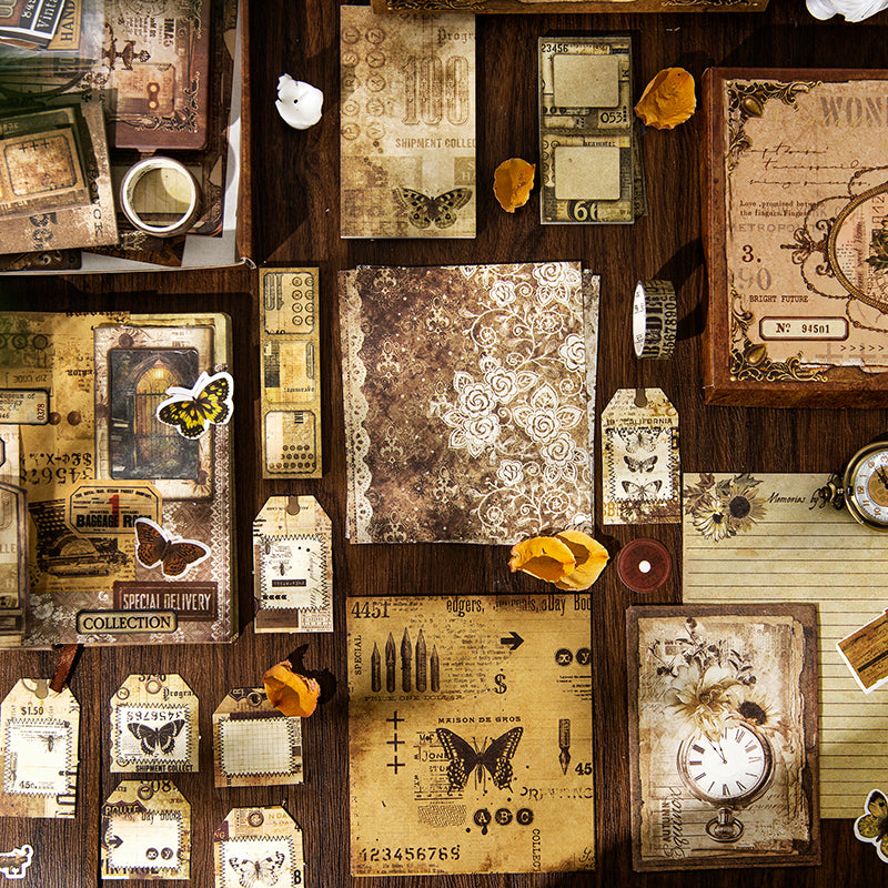 A rich flat lay showcasing the 153-piece vintage journaling kit contents. Features distressed background papers with delicate white lace patterns, butterfly washi stickers, vintage clock faces, and antique tags on a wooden desk.