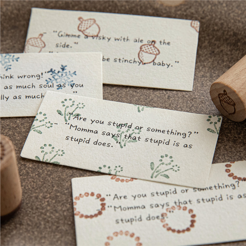 Stamped quote cards with acorn, snowflake, and wildflower borders, wooden stamps on grey surface.