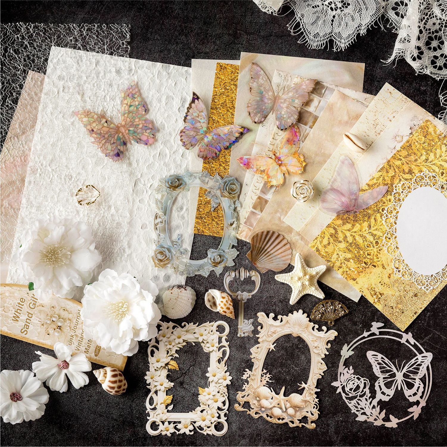 Flat lay of the Gilded Sands premium mixed media ephemera kit. Showcases elegant white 3D faux flowers, holographic PET butterflies, gold mesh, seashells, and intricate laser-cut frames on a dark background.