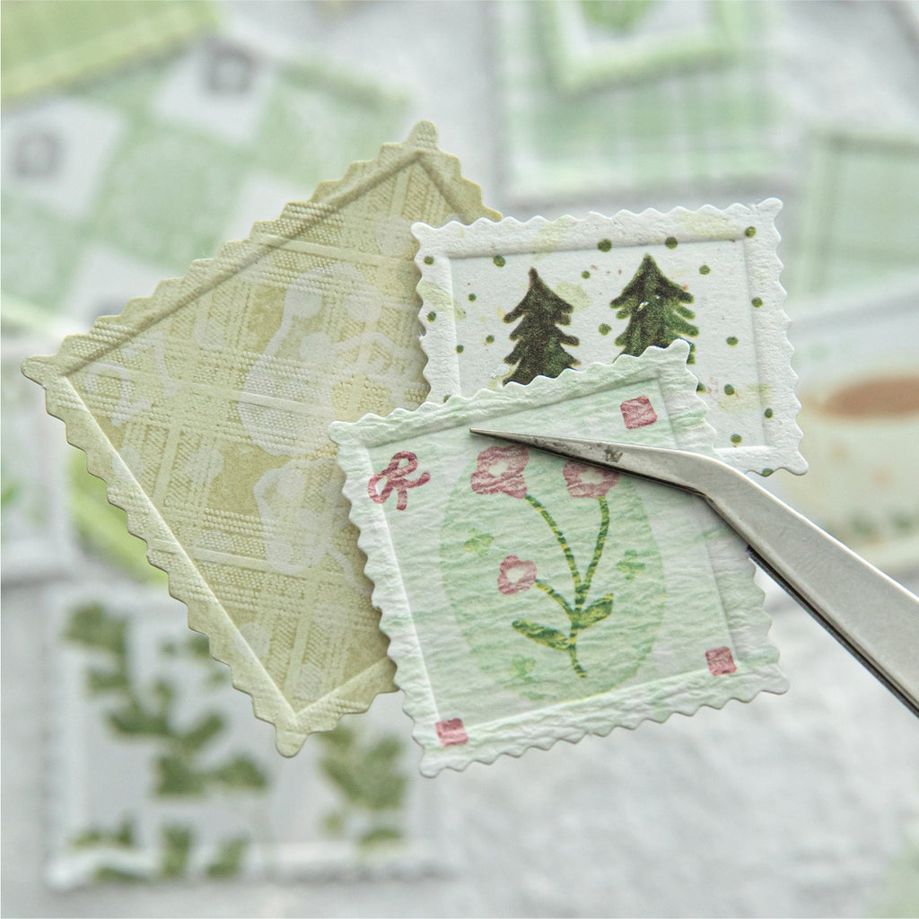 Macro shot using tweezers to hold a small green floral faux stamp from the Moss & Fern collection. Highlights the deep letterpress debossing, perforated postage edges, and premium textured specialty paper.