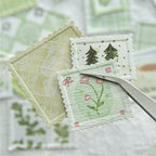 Macro shot using tweezers to hold a small green floral faux stamp from the Moss & Fern collection. Highlights the deep letterpress debossing, perforated postage edges, and premium textured specialty paper.