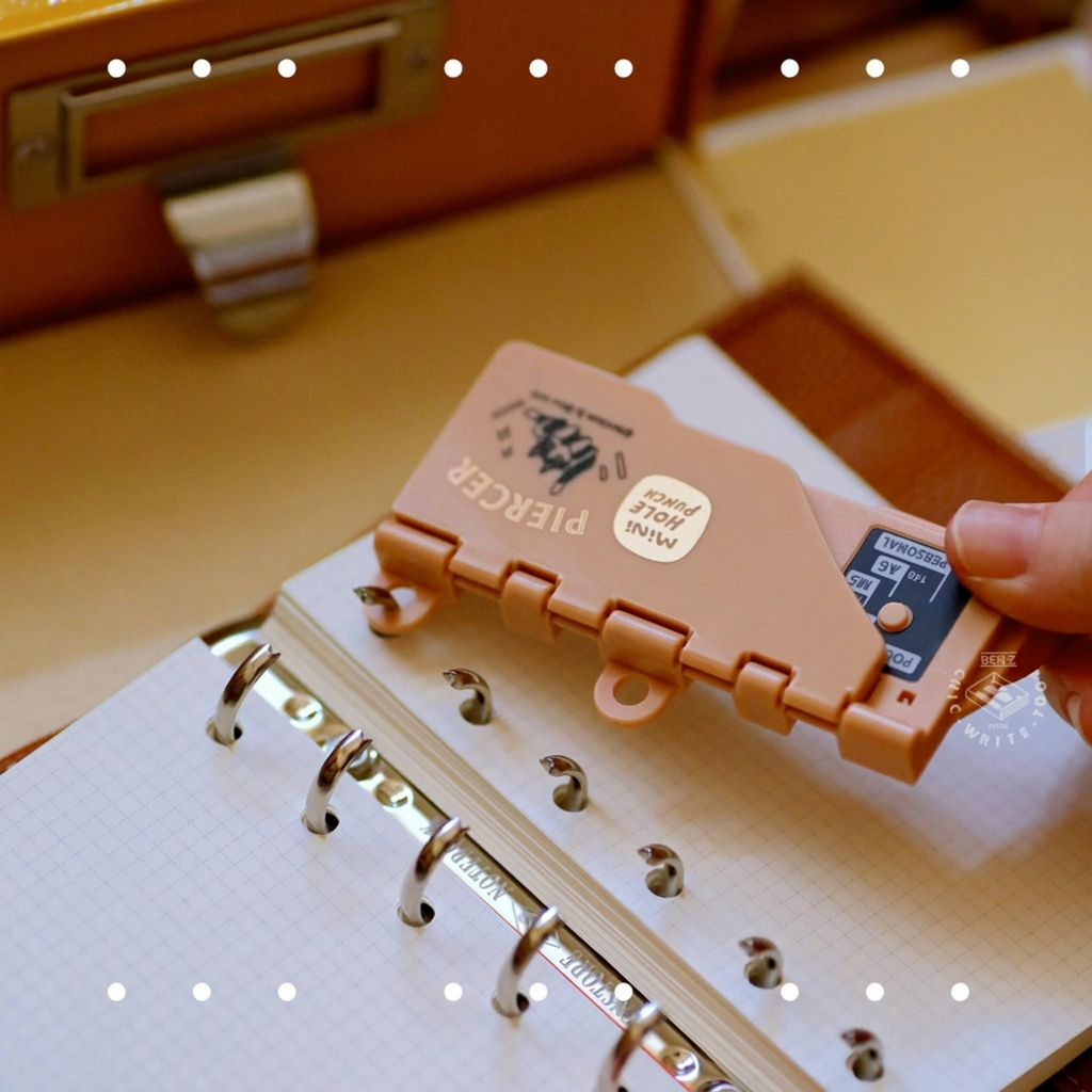 Hand holding brown portable 6-hole punch over white paper, multiple colored punches in background on desk.