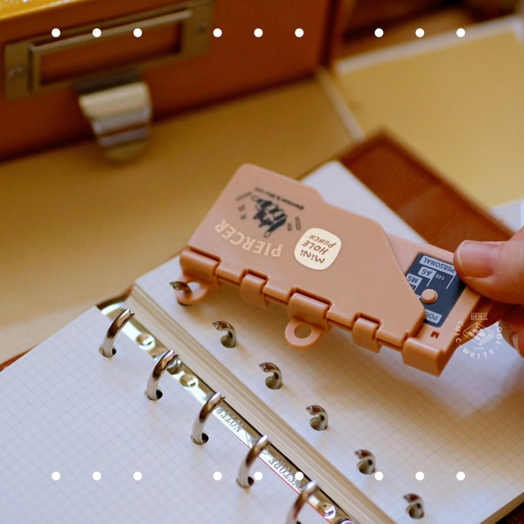 Hand holding brown portable 6-hole punch over white paper, multiple colored punches in background on desk.