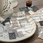 The Monochrome Archive style letterpress faux stamp ephemera scattered elegantly on a ceramic plate. Features black and grey designs like vintage keys, old houses, and coffee cups, styled with a cute panda figure.