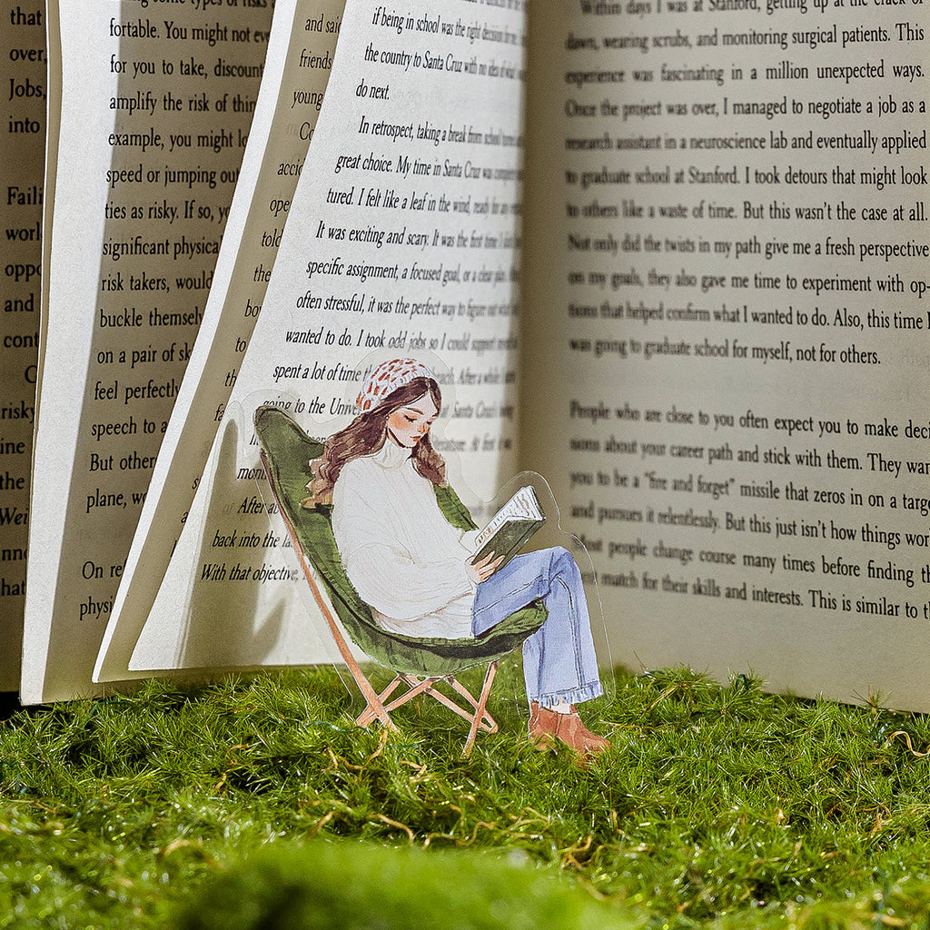 Green reading girl PET sticker clipped between open book pages on moss grass