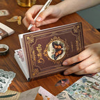 A person writing in the Nature's Whispers vintage brown butterfly journal. The A6 notebook is held over a wooden desk surrounded by aesthetic translucent butterfly stickers and classical ephemera for scrapbooking.