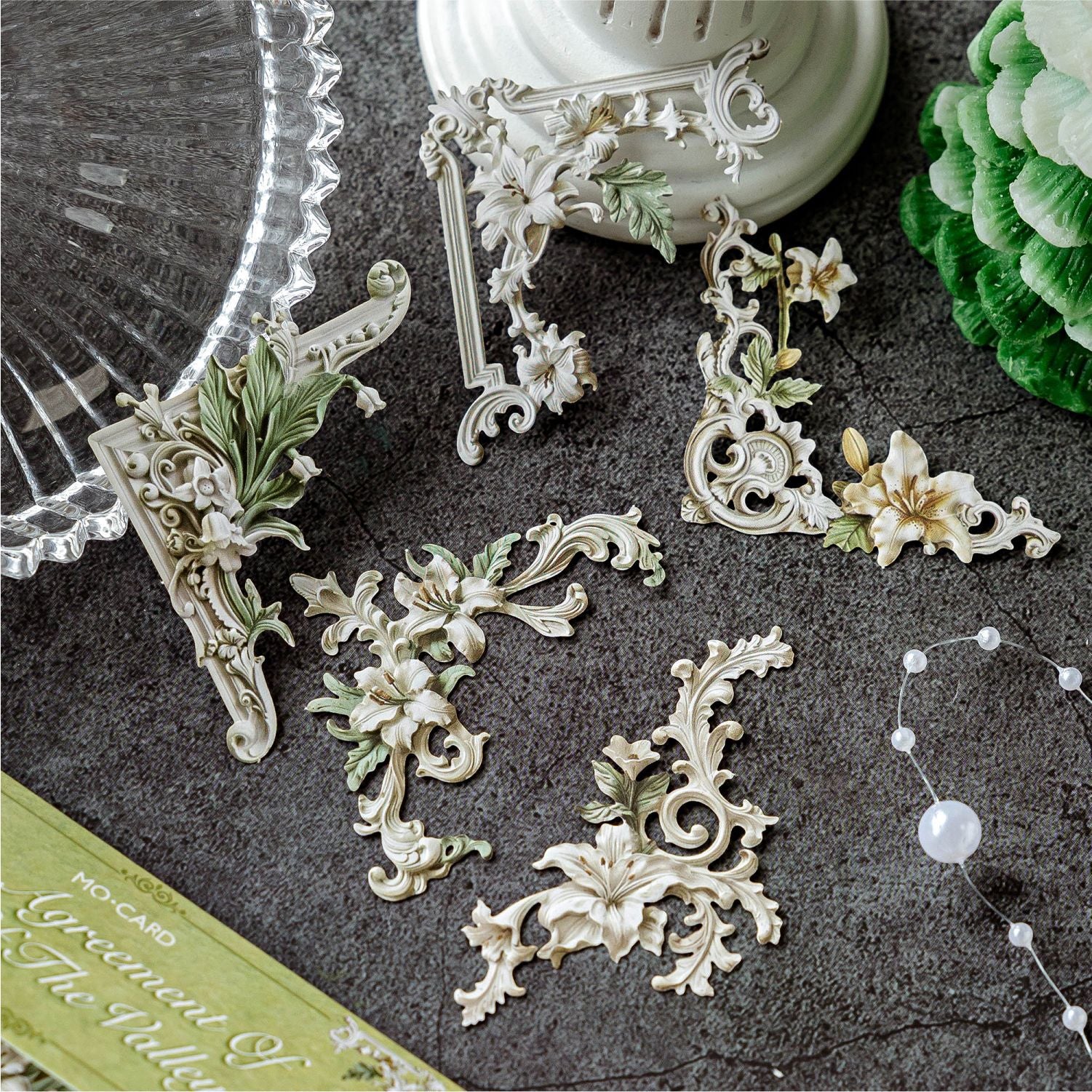 Close-up of the Secret Garden Lily style 3D relief corner frame ephemera. Showcases laser-cut vintage green pieces with lily of the valley and leaf motifs, arranged elegantly on a textured dark surface.