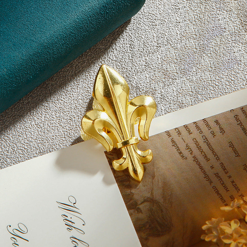 A single Luminous Gold Fleur-de-lis vintage metal journal clip clamping aged paper next to a quill and wax seal. Perfect Dark Academia aesthetic desk supply.