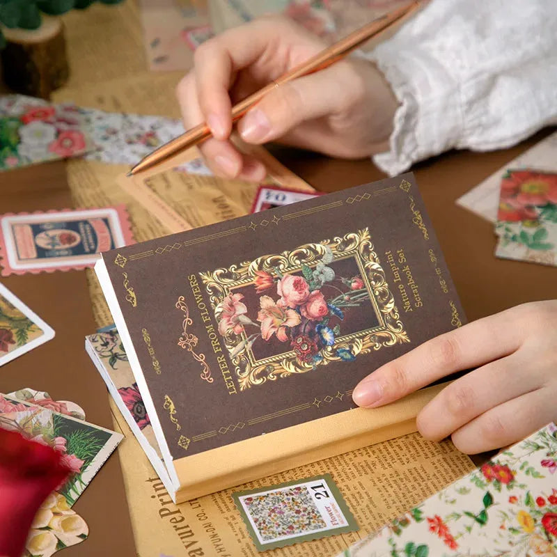 A detailed close-up of the "Letter From Flowers" journaling kit contents, focusing on semi-translucent PET botanical stickers with rose and floral patterns, small vintage music score ephemera cards, and a roll of washi tape next to the notebook.