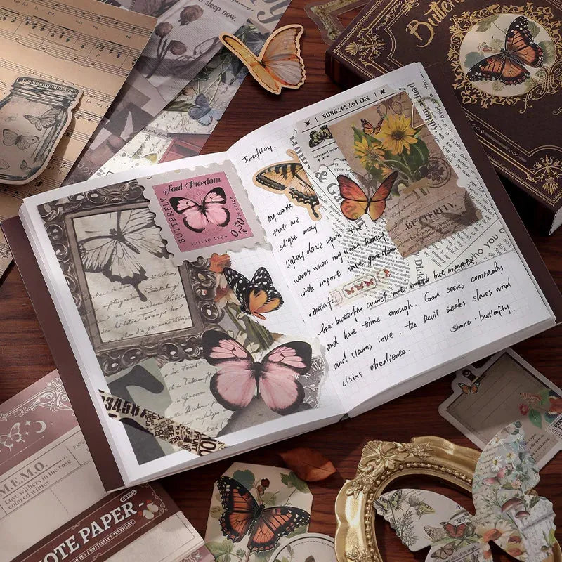 An open vintage notebook showcasing a beautifully layered junk journal spread. The pages are decorated with pink translucent butterfly PET stickers, vintage text papers, and botanical ephemera, surrounded by aesthetic scrapbooking supplies on a desk.