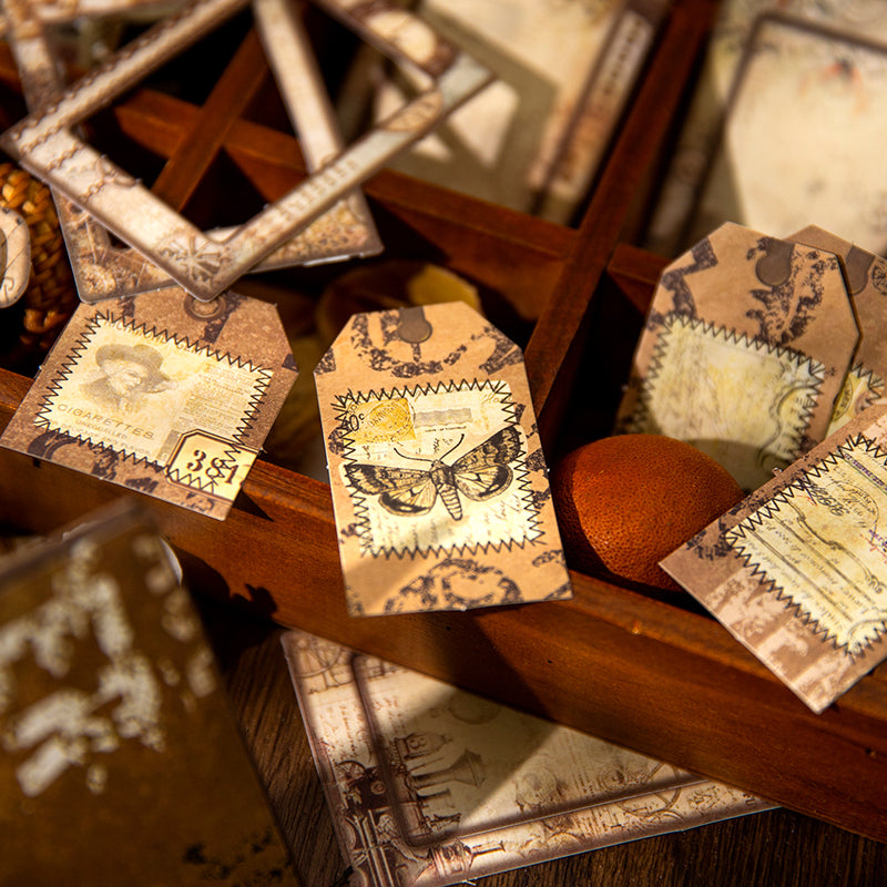 Detailed macro shot of vintage hang tags with faux stitched edges and beautiful butterfly illustrations, beautifully displayed in an antique wooden sorting tray for junk journaling.