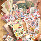 A hand holds the "Letter From Flowers" vintage floral notebook, highlighting its clear protective plastic cover. It rests on a sheet of scrapbooking paper alongside a pair of antique-style scissors, against a crafting desk background.