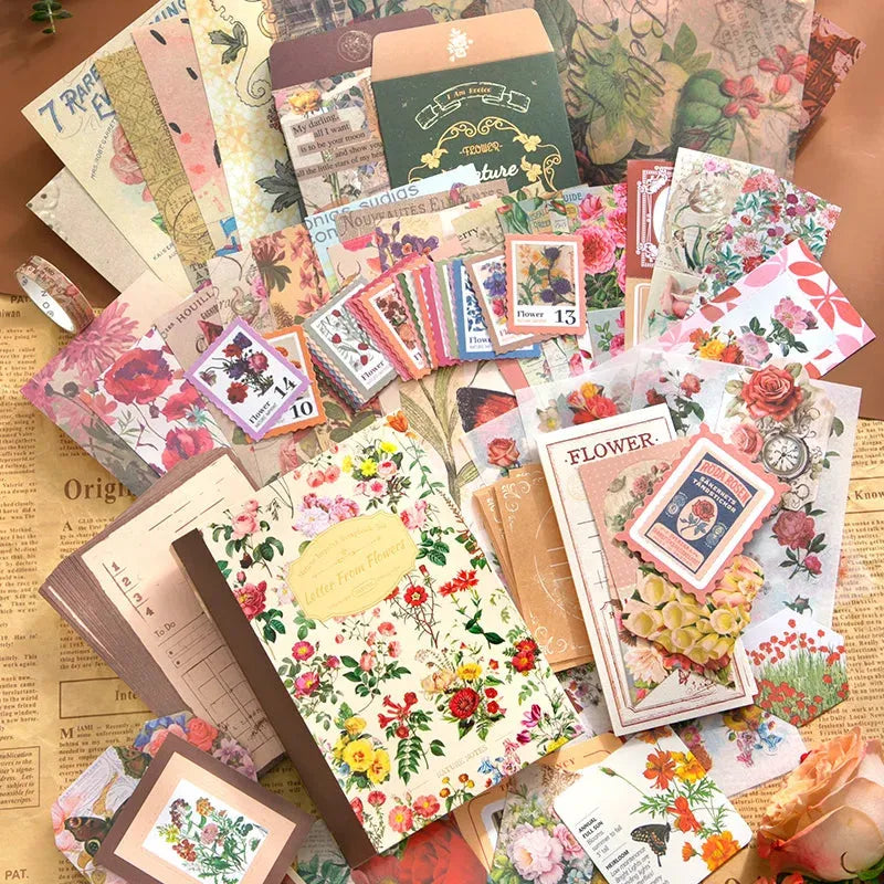 A hand holds the "Letter From Flowers" vintage floral notebook, highlighting its clear protective plastic cover. It rests on a sheet of scrapbooking paper alongside a pair of antique-style scissors, against a crafting desk background.