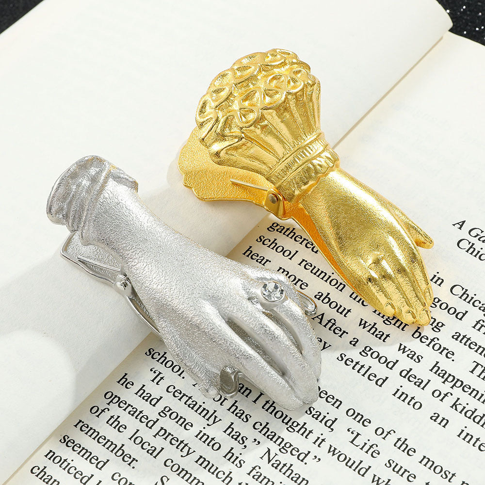 A close-up of two metal journal clips resting on an open book with printed text. The top clip is a large vintage gold hand holding a rose bouquet. The bottom clip is a small antique silver hand wearing a rhinestone ring. Both aesthetic tools are perfectly securing the book pages.