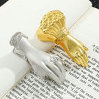 A close-up of two metal journal clips resting on an open book with printed text. The top clip is a large vintage gold hand holding a rose bouquet. The bottom clip is a small antique silver hand wearing a rhinestone ring. Both aesthetic tools are perfectly securing the book pages.