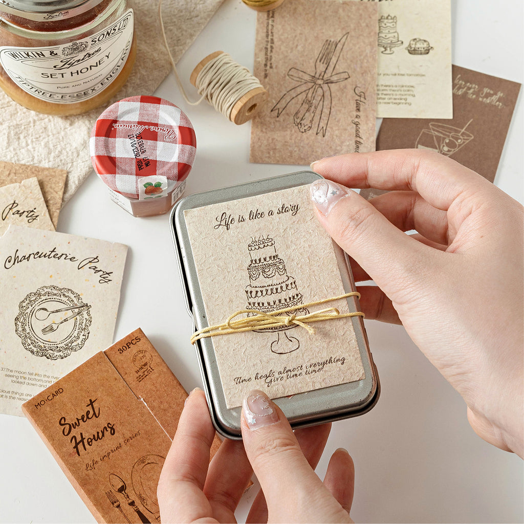 Hands holding vintage tin box with textured birthday cake journal card inside surrounded by kraft paper ephemera