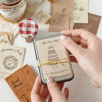 Hands holding vintage tin box with textured birthday cake journal card inside surrounded by kraft paper ephemera