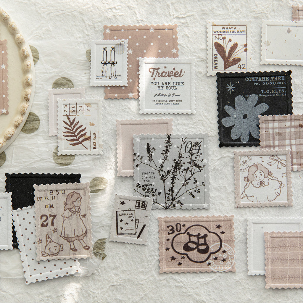 Aesthetic flat lay of vintage letterpress faux postage stamp ephemera scattered on white crumpled paper. Shows intricate textured details of floral, vintage girl, and typography designs from the mixed paper pack.