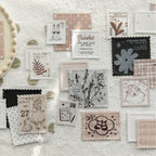 Aesthetic flat lay of vintage letterpress faux postage stamp ephemera scattered on white crumpled paper. Shows intricate textured details of floral, vintage girl, and typography designs from the mixed paper pack.
