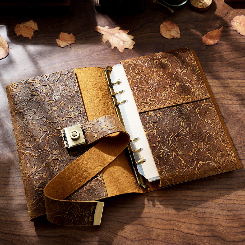 Top-down view of the Embossed Antique Floral leather journal lying completely open on a rustic wooden desk. Showcases the raw suede interior of the top-grain cowhide, the vintage metal lock strap, and the binder mechanism.